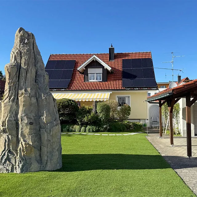 Haus mit Solarpaneelen auf dem Dach, davor zwei große künstliche Felsen auf einem gepflegten Rasen neben einer überdachten Einfahrt