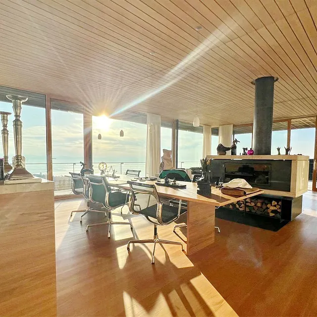 Helles, modernes Wohnzimmer mit großem Holztisch, mehreren Stühlen, Kamin und bodentiefen Fenstern mit Blick auf Wasser und Sonnenuntergang