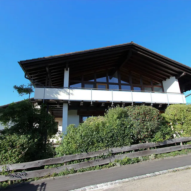 Zweistöckiges Haus mit großem Dachüberstand, Balkon und umgebender Bepflanzung an einer abschüssigen Straße