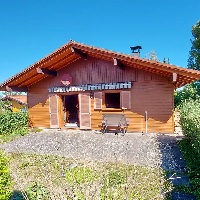 Einfamilienhaus mit Holzwand, offenem Fenster und offener Tür, davor eine Bank auf gepflastertem Hof, umgeben von grünen Büschen und Bäumen