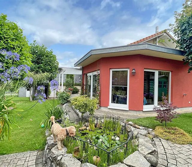 Garten mit kleinem Teich in einem steinernen Becken vor einem roten, eingeschossigen Haus mit großen Fenstern und umlaufender Terrasse