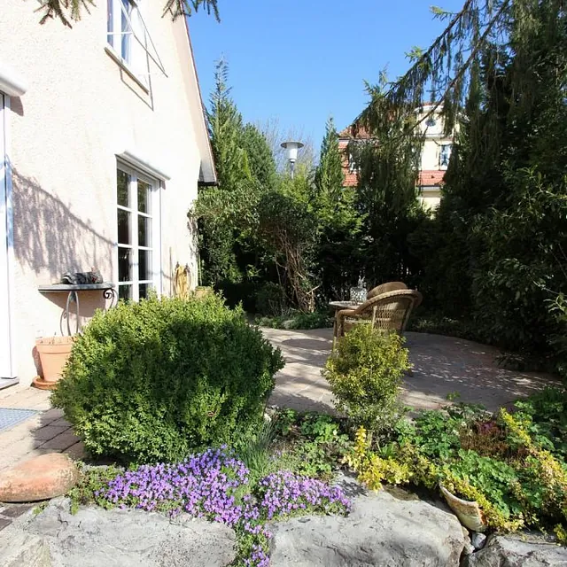  Ein Garten mit einer Steinmauer und einer einladenden Terrasse, umgeben von üppigem Grün und blühenden Pflanzen.