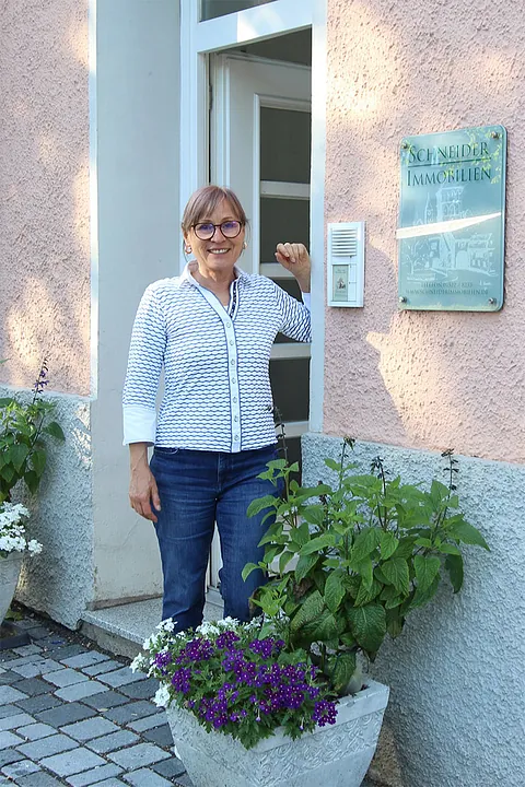 Frau mit Brille und weiß-blau gemusterter Bluse steht lächelnd vor einem Haus mit Schild 'Schneider Immobilien'
