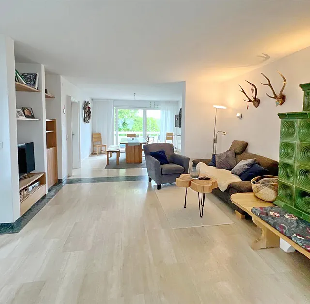 Großzügiges Wohnzimmer mit grünem Kachelofen, Sitzbank mit gemustertem Polster, Sesseln, Holztisch und Hirschgeweihen an der Wand