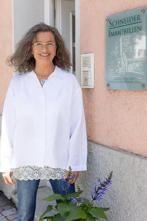 Frau mit Brille und weißer Bluse steht lächelnd vor einem Haus neben einem Schild mit der Aufschrift 'Schneider Immobilien'