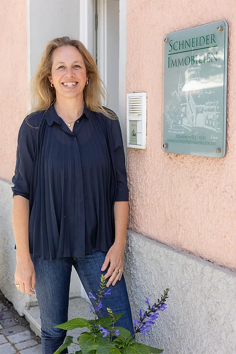 Frau mit langem blondem Haar, dunkelblauer Bluse und Jeans steht lächelnd vor einem Haus mit Schild 'Schneider Immobilien'