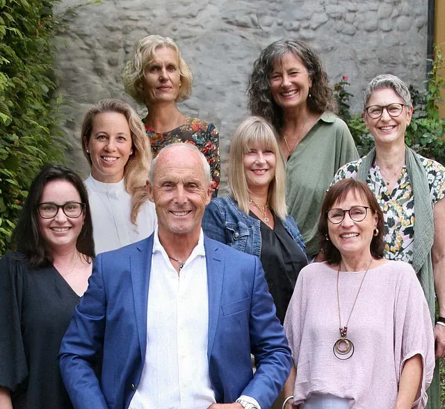 Gruppe von acht Personen, sechs Frauen und ein Mann, stehen vor einer Steinmauer und grünen Pflanzen, alle lächeln in die Kamera