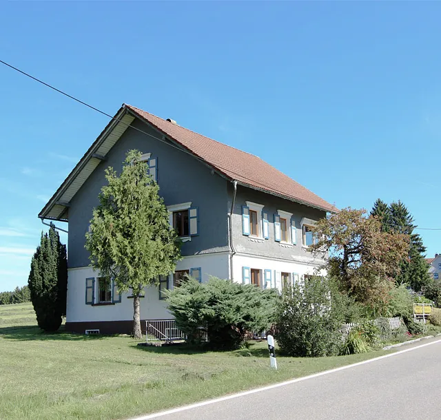 Zweistöckiges Haus mit Satteldach an einer Landstraße umgeben von Bäumen und Wiesen
