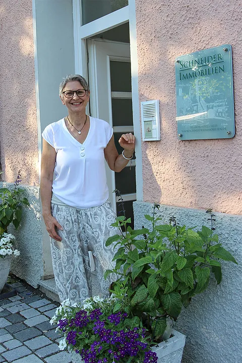 Eine lächelnde Frau mit Brille und weißer Bluse steht vor einem Hauseingang neben einem Schild mit der Aufschrift 'Schneider Immobilien' und mehreren Blumentöpfen.