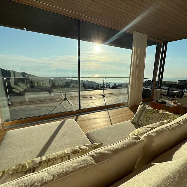 Wohnzimmer mit hellbeiger Couch vor bodentiefen Fenstern und Blick auf eine Terrasse und eine Landschaft mit Hügeln im Sonnenlicht