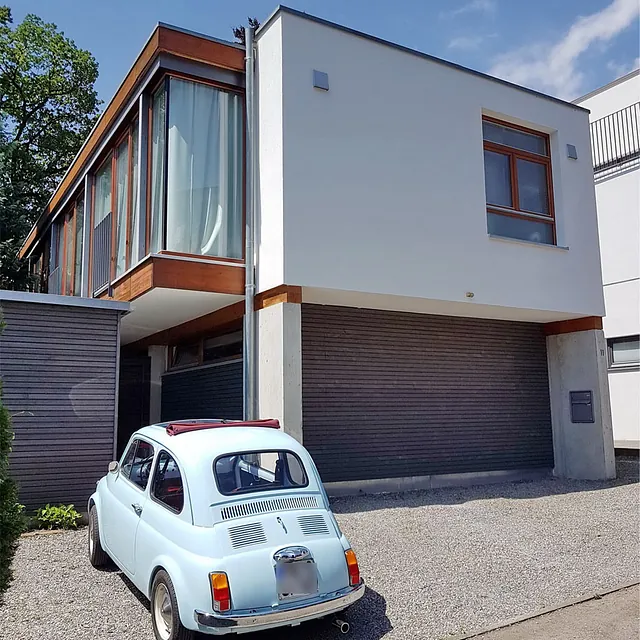 Kleines hellblaues Auto steht vor einem modernen zweistöckigen Haus mit großen Fenstern und weißer Fassade